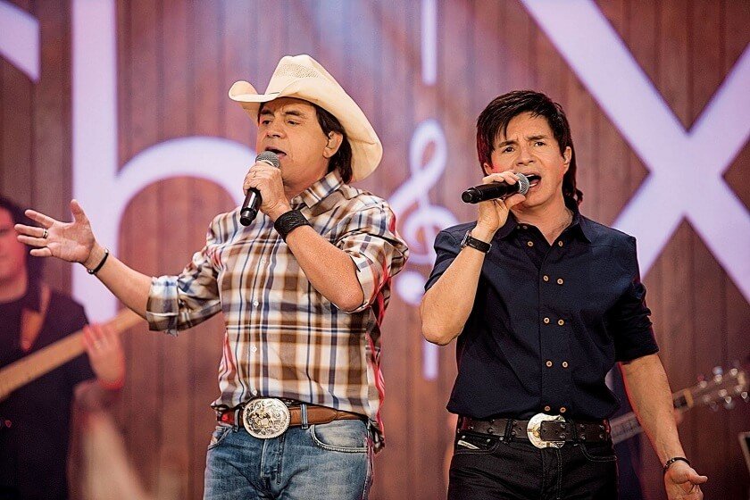 Foto colorida da dupla Chitãozinho e Xororó. Os artistas estão no palco, cantando - Metrópoles