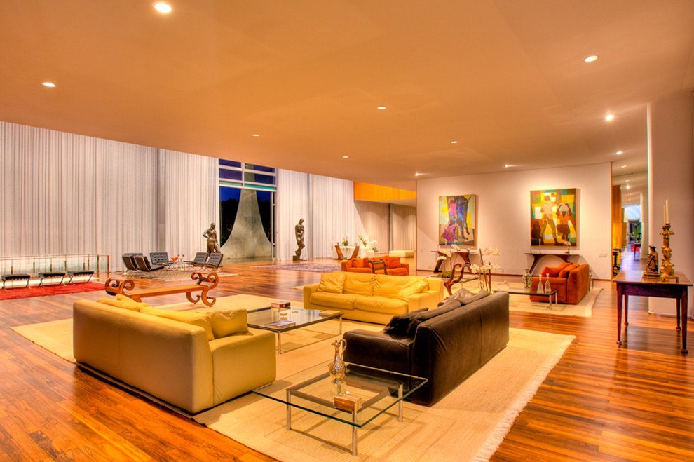 Foto colorida de uma sala com dois sofás amarelos, um preto e um tapete branco. Além de cortinas brancas