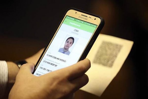 Fotografia colorida de pessoa segurando um celular com a mão e a cnh física na outra mão. No celular aparece a imagem da carteira digital e na cnh o qr code para acesso ao documento