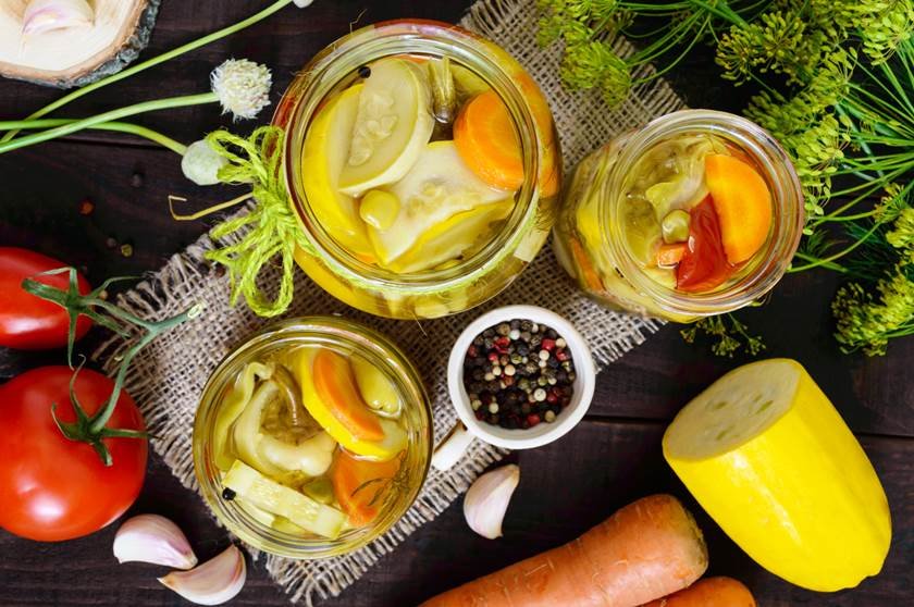 Pickles: vegetable assortment (zucchini, pepper, carrots, tomato, green peas) in glass jars on a dark wooden background. The top view