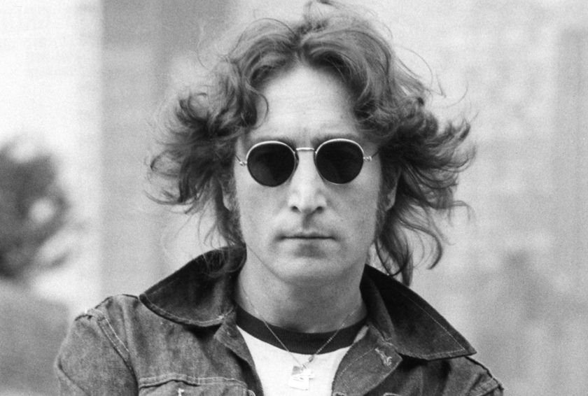 Foto de John Lennon em preto e branco. Homem de pele clara, com óculos escuros, cabelo longo na altura do ombro, jaqueta e blusa branca - Metrópoles