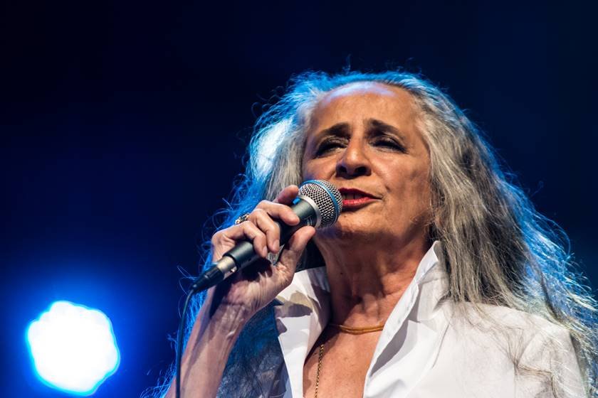 Foto colorida da cantora Maria Bethânia. Ela segura um microfone com a mão direita e usa uma blusa branca. Tem cabelos grisalhos - Metrópoles