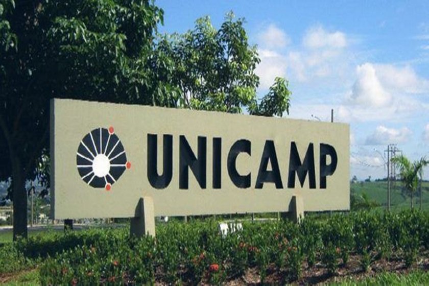 Placa da Unicamp. O objeto é feito de concreto é mostra a logo da universidade seguida pela sigla. Ela está cercada de plantas | Metrópoles