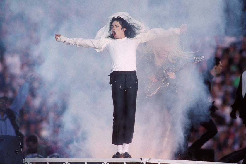 Foto colorida de Michael Jackson. Ele está em um palco com os braços abertos - Metrópoles