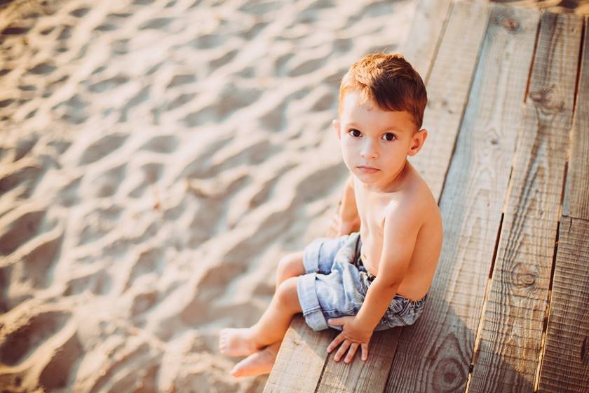 The theme is a child and summer beach vacation. A small Caucasian boy sits sideways on a wooden pier and looks at the camera on a sandy beach and a pond, a river. With bare legs in blue denim shorts