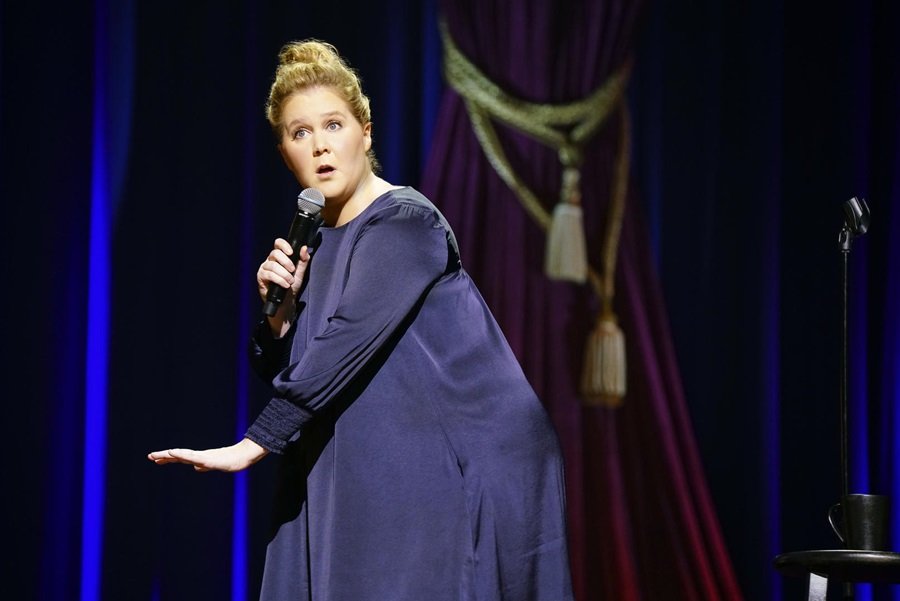 Foto da atriz Amy Schumer em um show. Ela está no palco, de vestido azul e microfone na mão - Metrópoles
