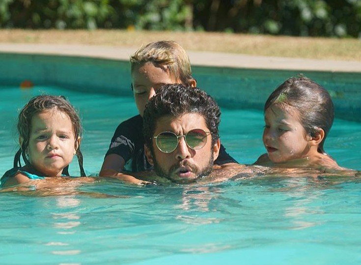 Foto colorida de quatro pessoas em uma piscina: um homem, dois meninos e uma menina - Metrópoles