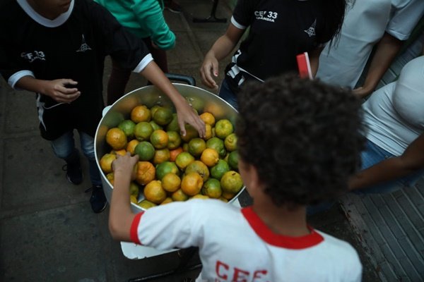 Menino negro com cabelo cacheado de costas pegando laranja em panela grande cinza. Outras pessoas também pegam laranja na panela
