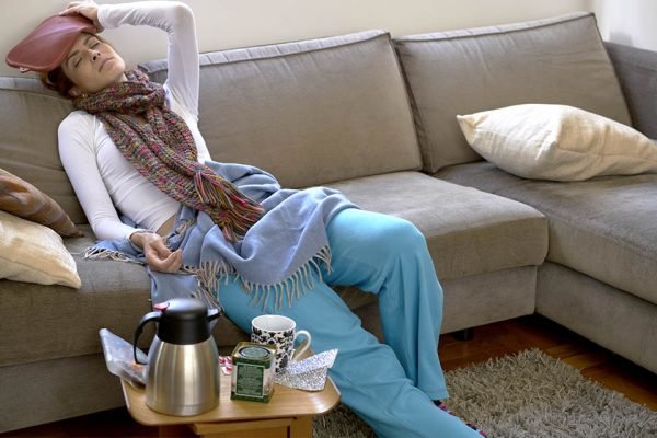 Fotografia colorida. Mulher sentada no sofá com a mão na cabeça e roupa de frio