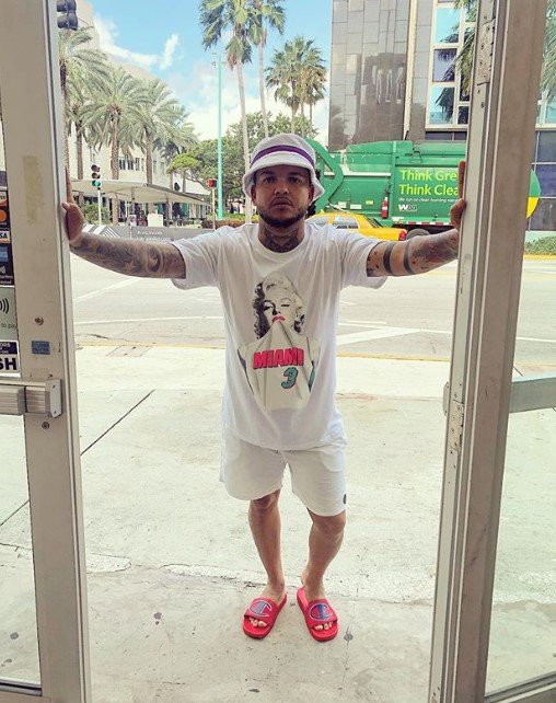 Foto do cantor Suel. Ele usa chinelo rosa, short branco e camisa branca. Está com uma viseira e se apoia com as duas mãos em uma porta. - Metrópoles