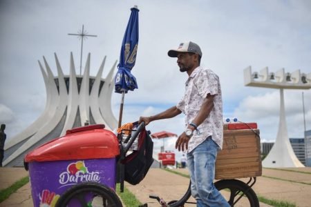 Homem de boné em pé ao lado de carrinho de sorvete. Ao fundo está a Catedral de Brasília