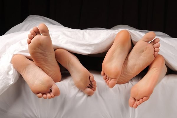 Foto colorida de seis pés saindo debaixo de um edredom branco deitados juntos em uma cama - Metrópoles