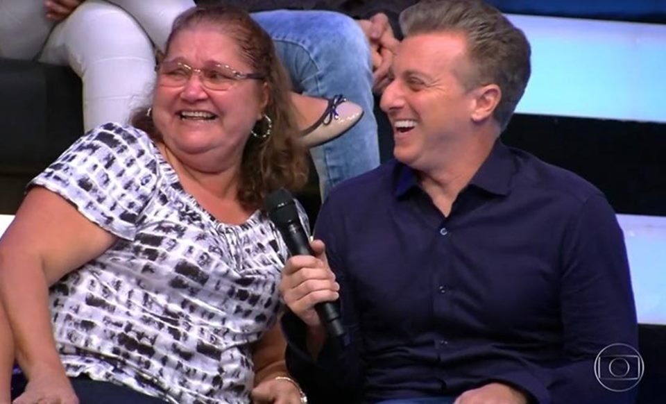 Luciano Huck aponta microfone para mulher na plateia e ambos riem