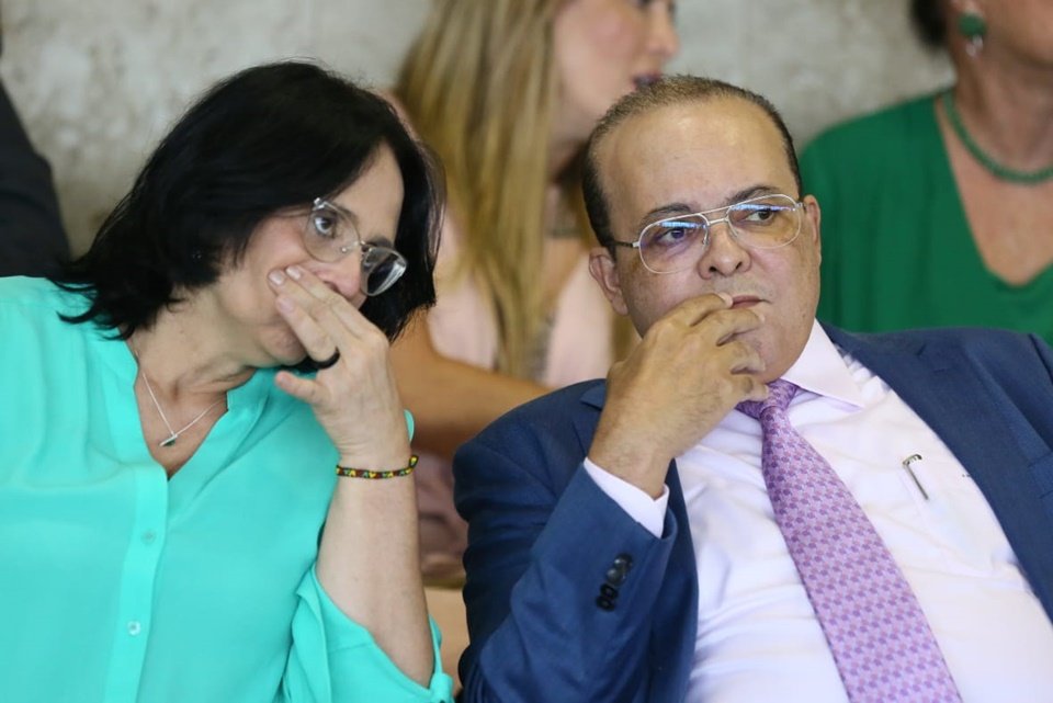 Fotografia colorida mostra mulher de azul com mão na boca e homem de terno ao lado