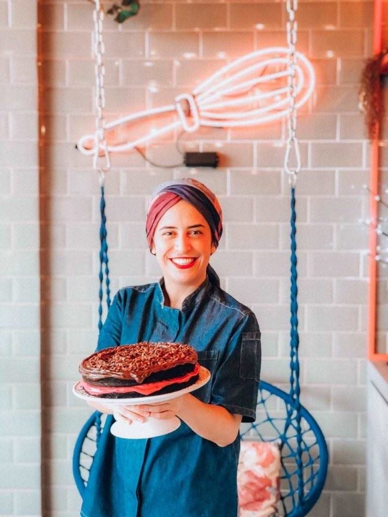 A chef Jamile Bilu, da confeitaria Bem Delicioso, segura um bolo em frente a um balanço azul e um fuê em neon