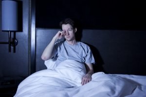 Imagem de homem com insônia sentado na cama à noite coçando os olhos - Metrópoles