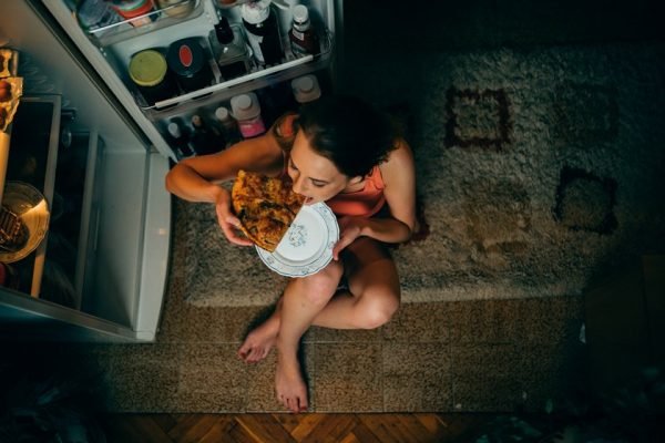 Mulher come pizza em frente à geladeira