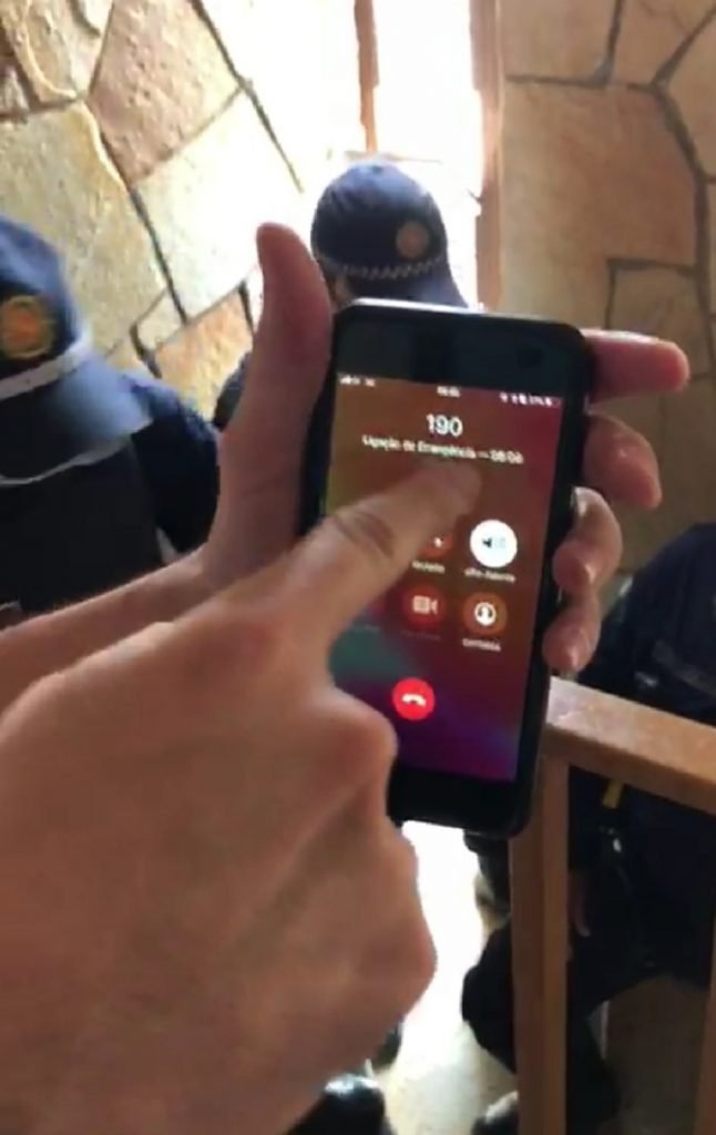 Mão segurando celular ligando para 190 e PMs ao fundo