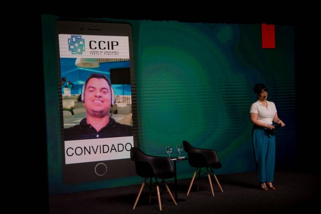 Mulher ao lado de cadeiras vazias e um telão com imagem de homem