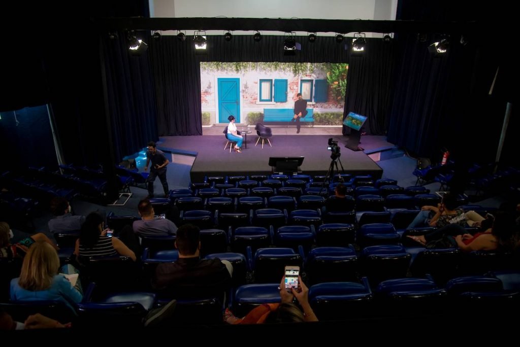auditorio com plateia assistindo e uma mulher falando no palco