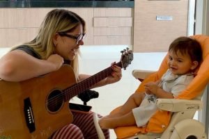 Imagem da cantora marília mendonça com um violão, brincando com o filho Leo, que está sentado em uma cadeirinha - Metrópoles