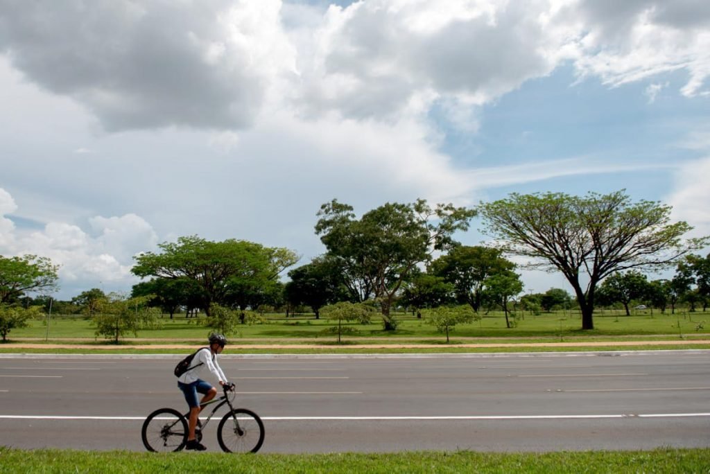 Cicilista em bicicleta passando em pista; grama e árvores ao fundo