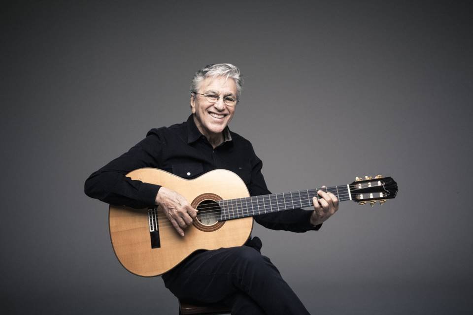 Foto colorida de Caetano Veloso. Ele está de roupa preta, usa óculos de grau e tem cabelos grisalhos. Ele segura um violão - Metrópoles