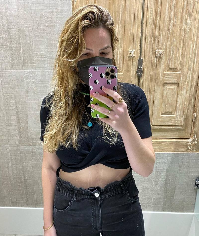 Foto colorida de uma mulher loira, tirando uma selfie no espelho, usando blusa preta e calça preta - Metrópoles