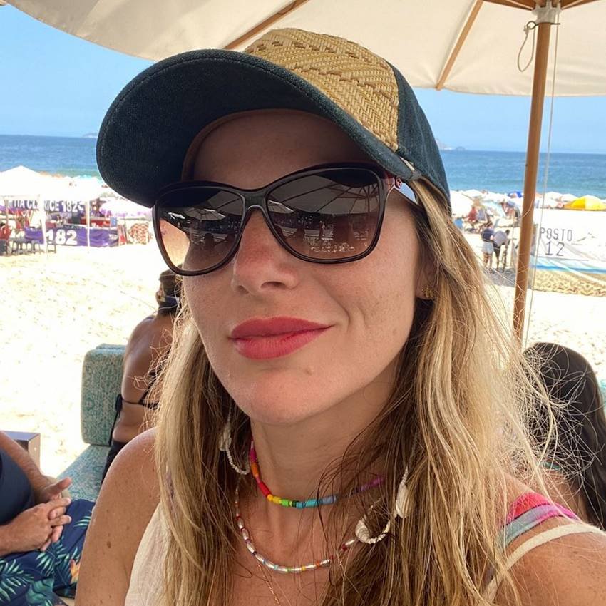 Foto colorida de uma mulher loira, usando óculos de sol e boné, na praia - Metrópoles