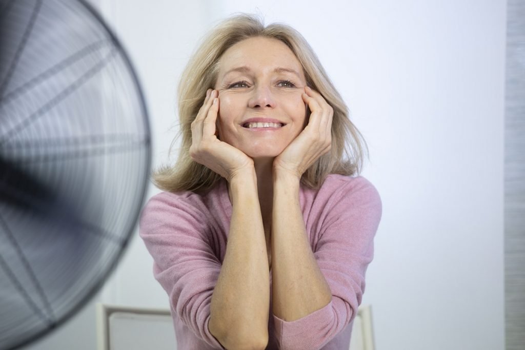 Imagem colorida: mulher de meia idade sentada em frente a ventilador - Metrópoles