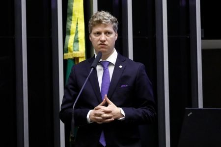 O deputado do partido Novo pelo Rio Grande do Sul, Marcel van Hattem, discursa no plenário da Câmara dos Deputados diante de microfone. Ele está de mãos cruzadas - Metrópoles