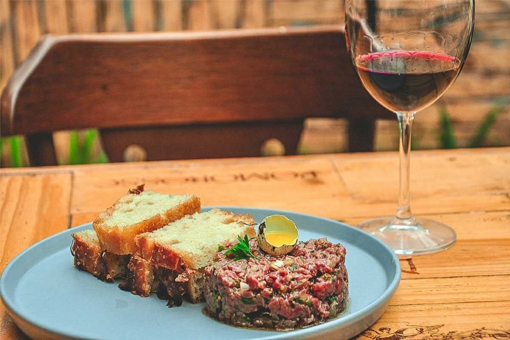 Prato com steak tartare e torradas e taça de vinho, tudo sobre uma mesa de madeira