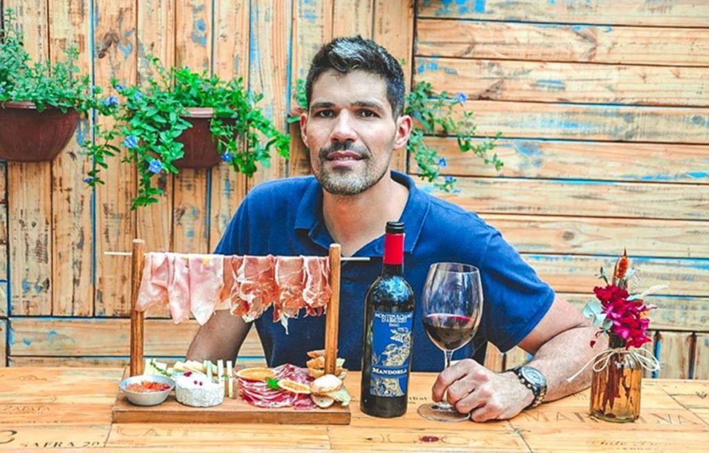 Homem segurando uma taça de vinho que está sobre uma mesa de madeira junto com uma garrafa de vinho e uma tábua de frios