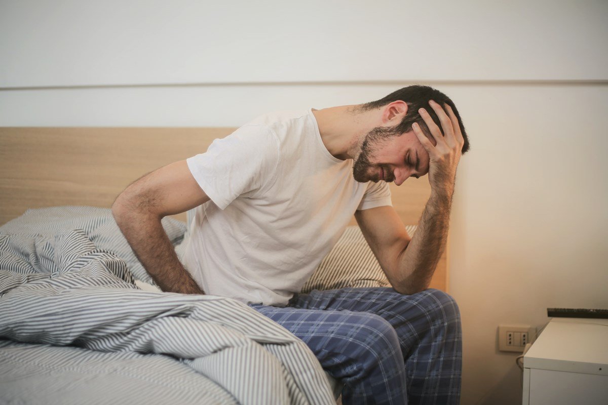 Foto colorida de homem sentado na cama com dor de cabeça de ressaca