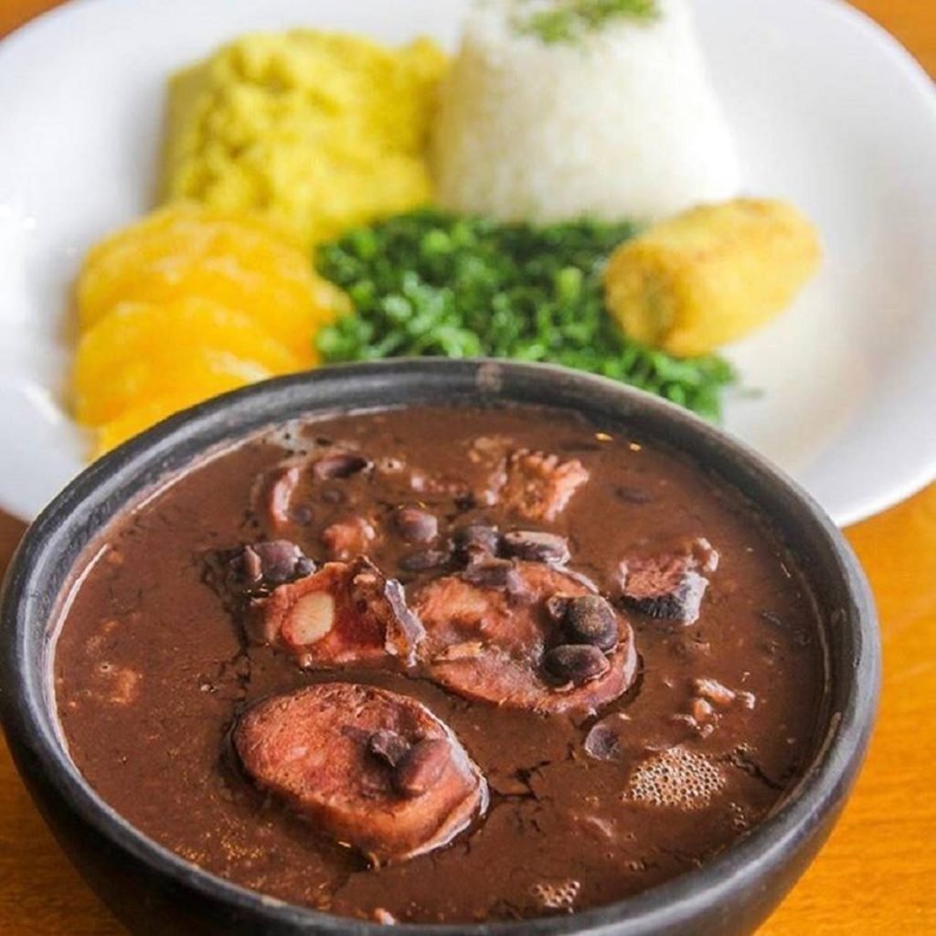 Panela com feijoada e prato com arroz, couve, farofa e laranja