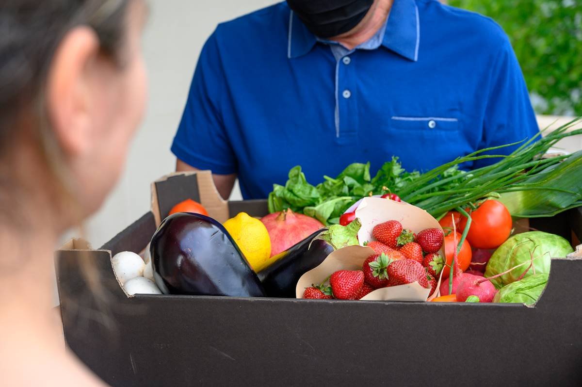 Homem entrega caixa com frutas e verduras para mulher que aparece apenas no canto da imagem