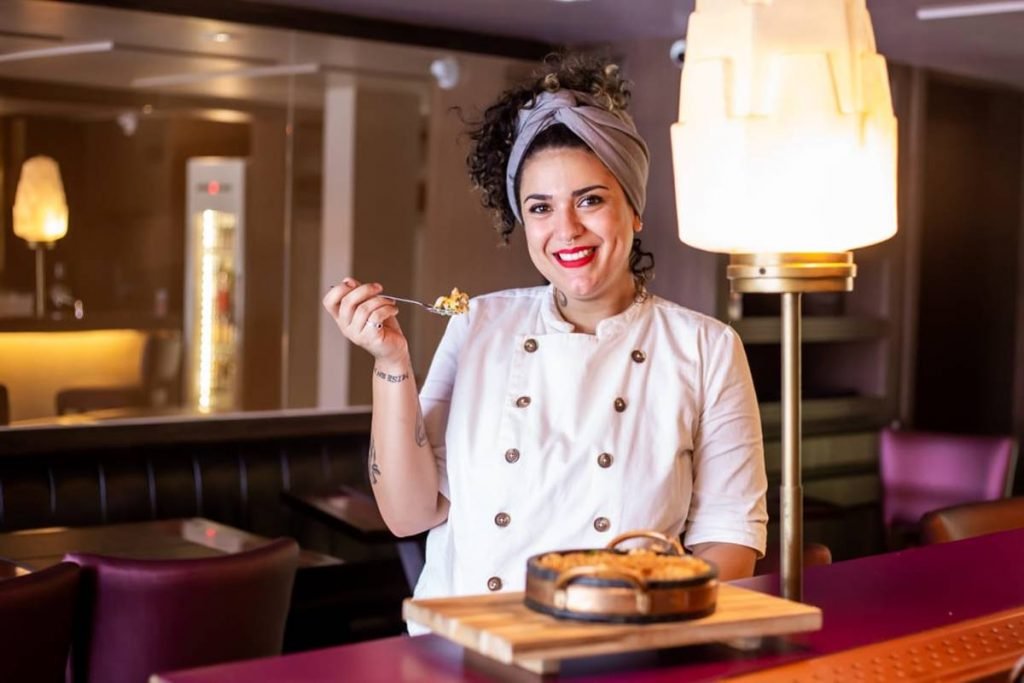 Mulher com cabelos cacheados vestida com uma dolmã de chef e segurando um garfo com comida