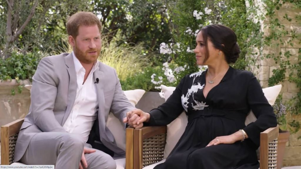 Príncipe Harry e Meghan Markle sentados em cadeiras de mãos dadas