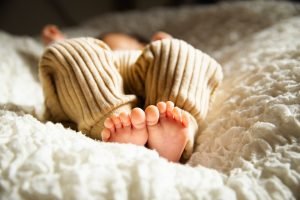 Foto mostra os pés de bebê vestido em roupa bege em primeiro plano presa