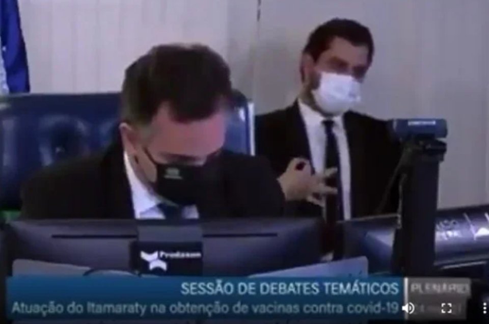 Homem de preto ao fundo, de máscara branca e terno fazendo gesto de ok