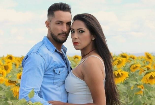 Homem com camisa azul e mulher com blusa branca se abraçando com girassóis atrás