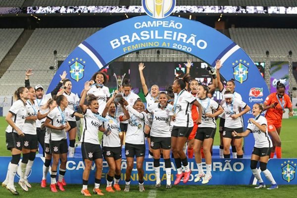 Imagem colorida de time feminino do Corinthians em frente a pódio de campeãs do Brasileirão Feminino de 2022 - Metrópoles