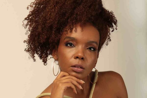 Foto colorida de Thelma Assis com a mão apoiada no queixo e cabelo em frente ao rosto - Metrópoles