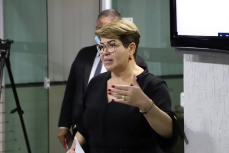Mulher loira e branca vestida de preto com uma expressão de fala e uma das mãos levantadas