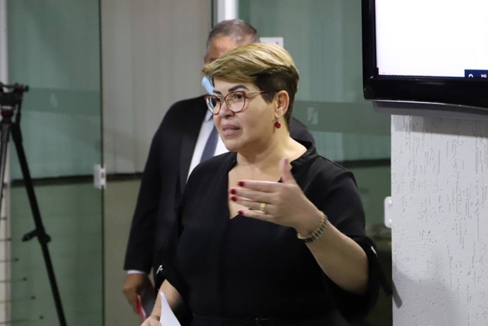 Mulher loira e branca vestida de preto com uma expressão de fala e uma das mãos levantadas