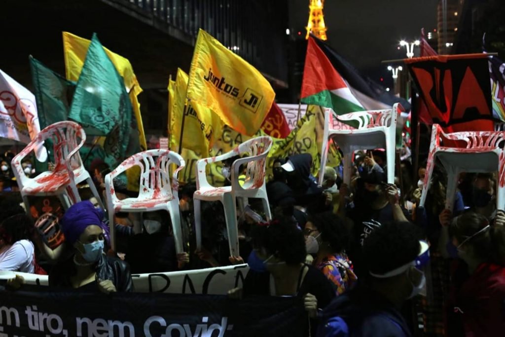 Manifestantes caminham com faixas, bandeiras e cadeiras com manchas vermelhas