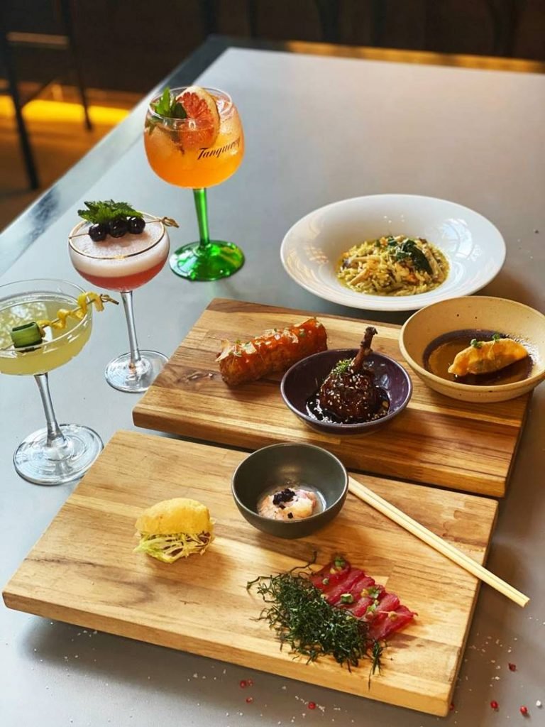Tábuas de madeira com pequenas porções de comida, um prato de risoni ao fundo e drinques na lateral
