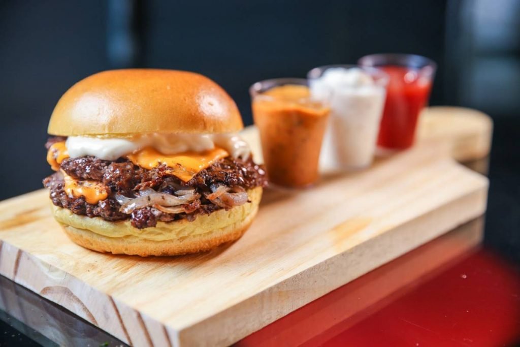 Hambúrguer com pão, carne e queijo em cima de tábua com maioneses ao lado