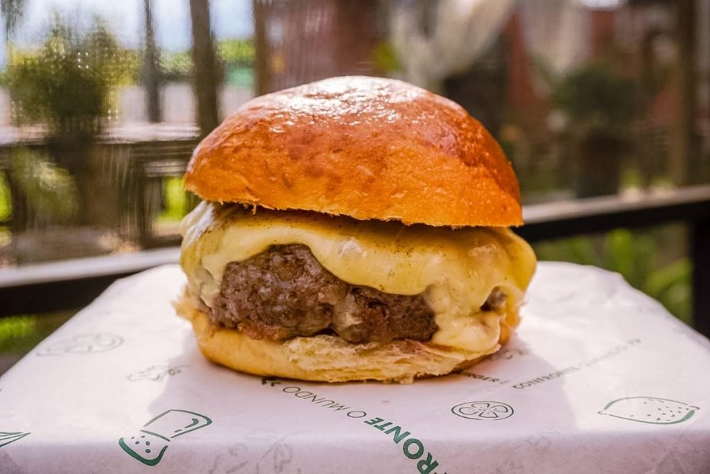 Hambúrguer com pão, carne e queijo em cima de tábua com jardim ao fundo