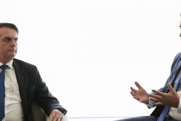 Jair Bolsonaro e Ciro Nogueira conversam sentados em fundo branco. Ciro fala e gesticula, olhando para frente, e Bolsonaro o observa - Metrópoles